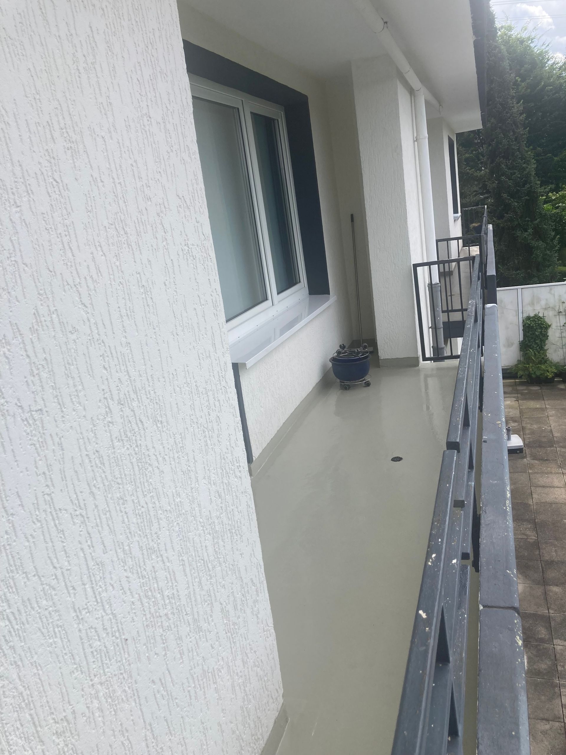 Außenansicht eines Balkons mit weißen strukturierten Wänden, einem Fenster und einem schwarzen Geländer.