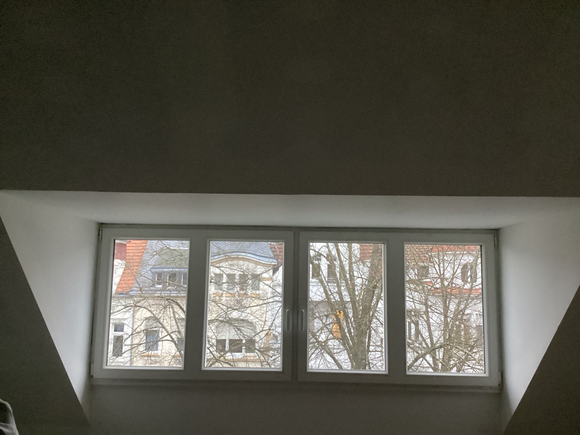 Fenster in einem Dachgeschoss mit Blick auf Gebäude und Bäume, eingerahmt von weißen Wänden und einer weißen Decke.