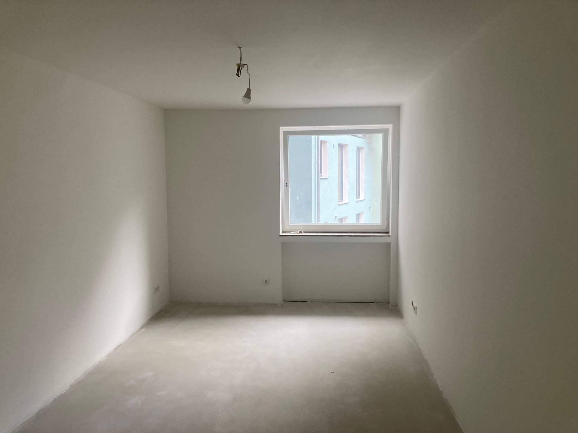 Leerer, frisch gestrichener Raum mit Fenster und Beleuchtung. Durch das Fenster ist ein hellblaues Gebäude zu sehen.