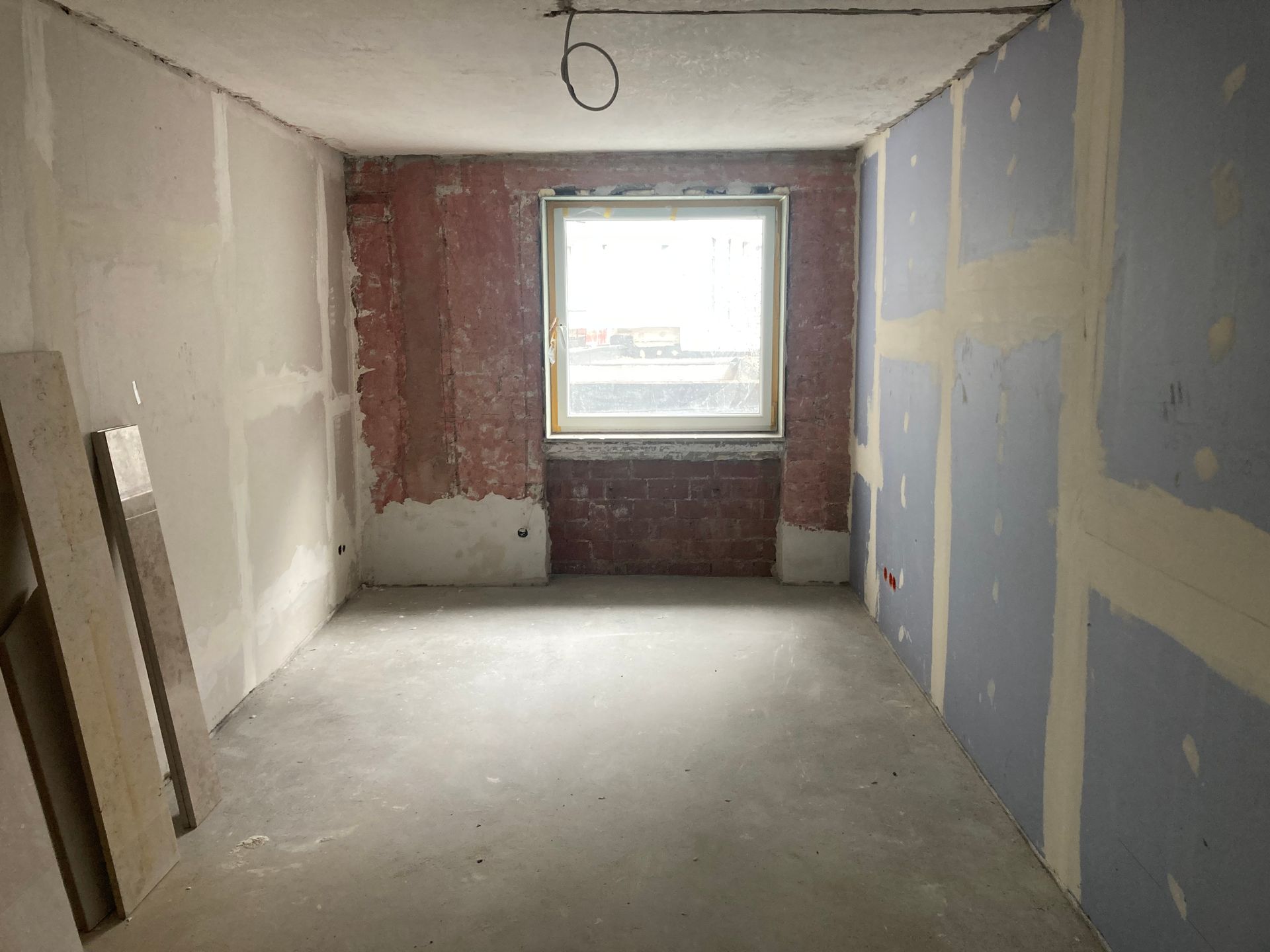 Unfertiger Raum mit Trockenbauwänden und Betonboden, Fenster in der Mitte.