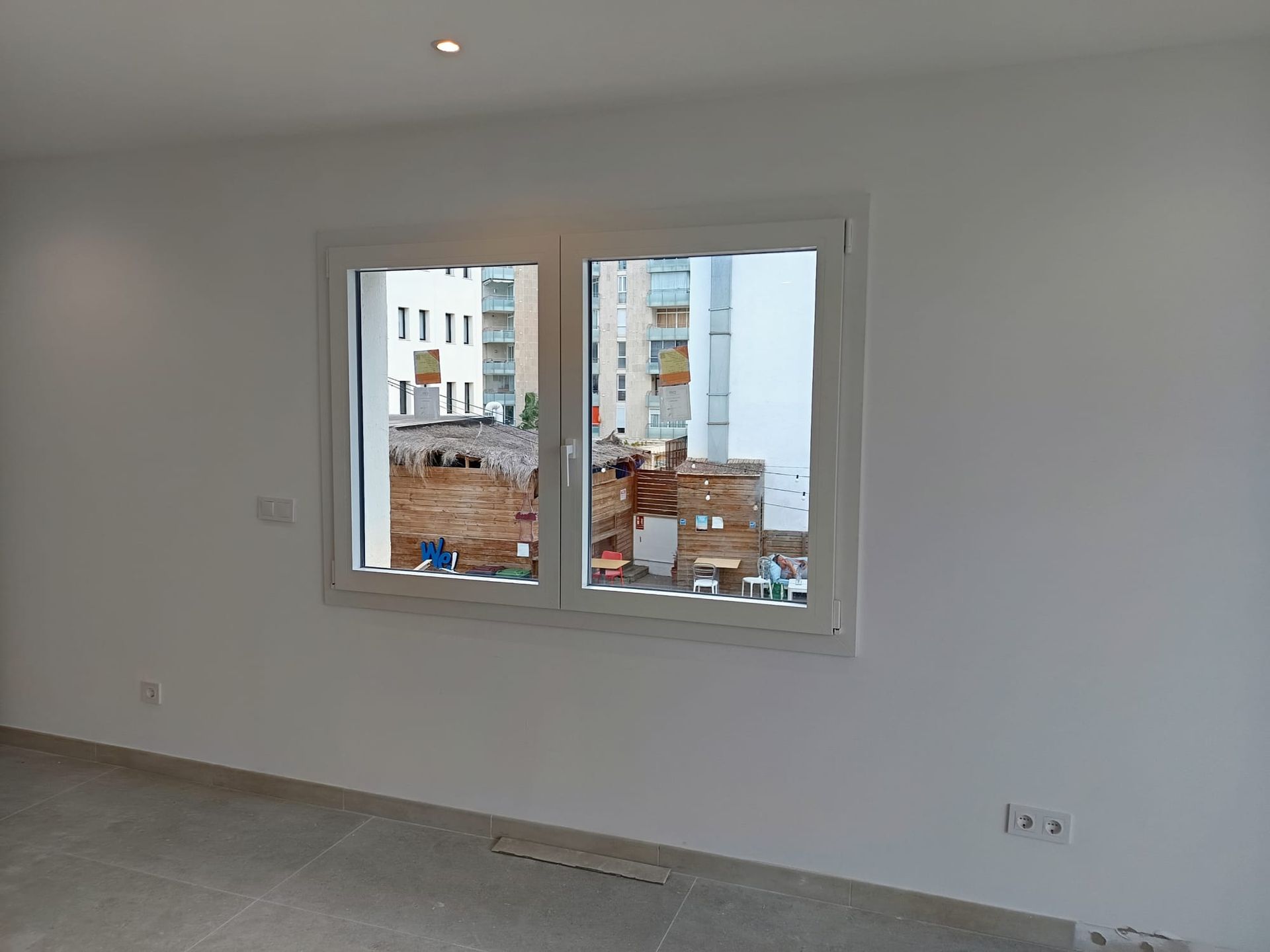 Habitación interior con una ventana con marco blanco. Vista de otros edificios, paredes blancas y suelo gris.