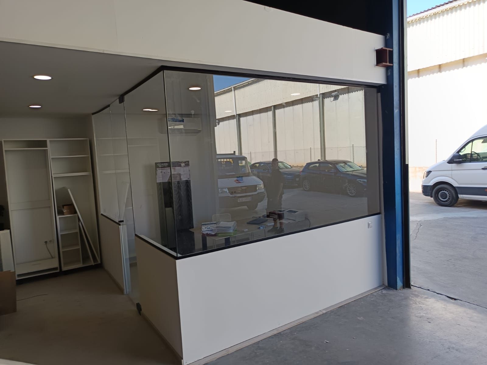 Un local comercial con una gran ventana y muebles blancos; una furgoneta blanca está estacionada afuera.