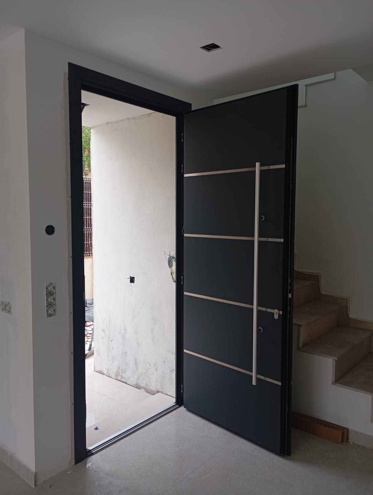 Puerta negra moderna y abierta con tirador plateado y detalles horizontales. La entrada da al exterior.