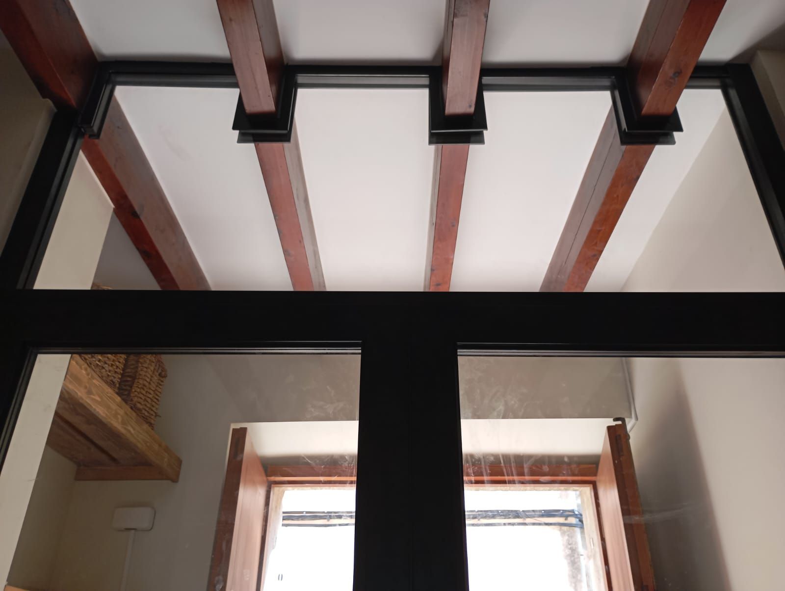Vista interior: vigas de madera en el techo sostenidas por soportes de metal negro sobre una ventana.