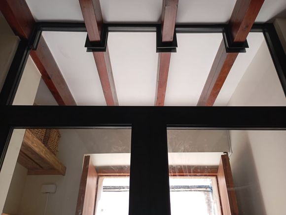 Vista interior: vigas de madera en el techo sostenidas por soportes de metal negro sobre una ventana.