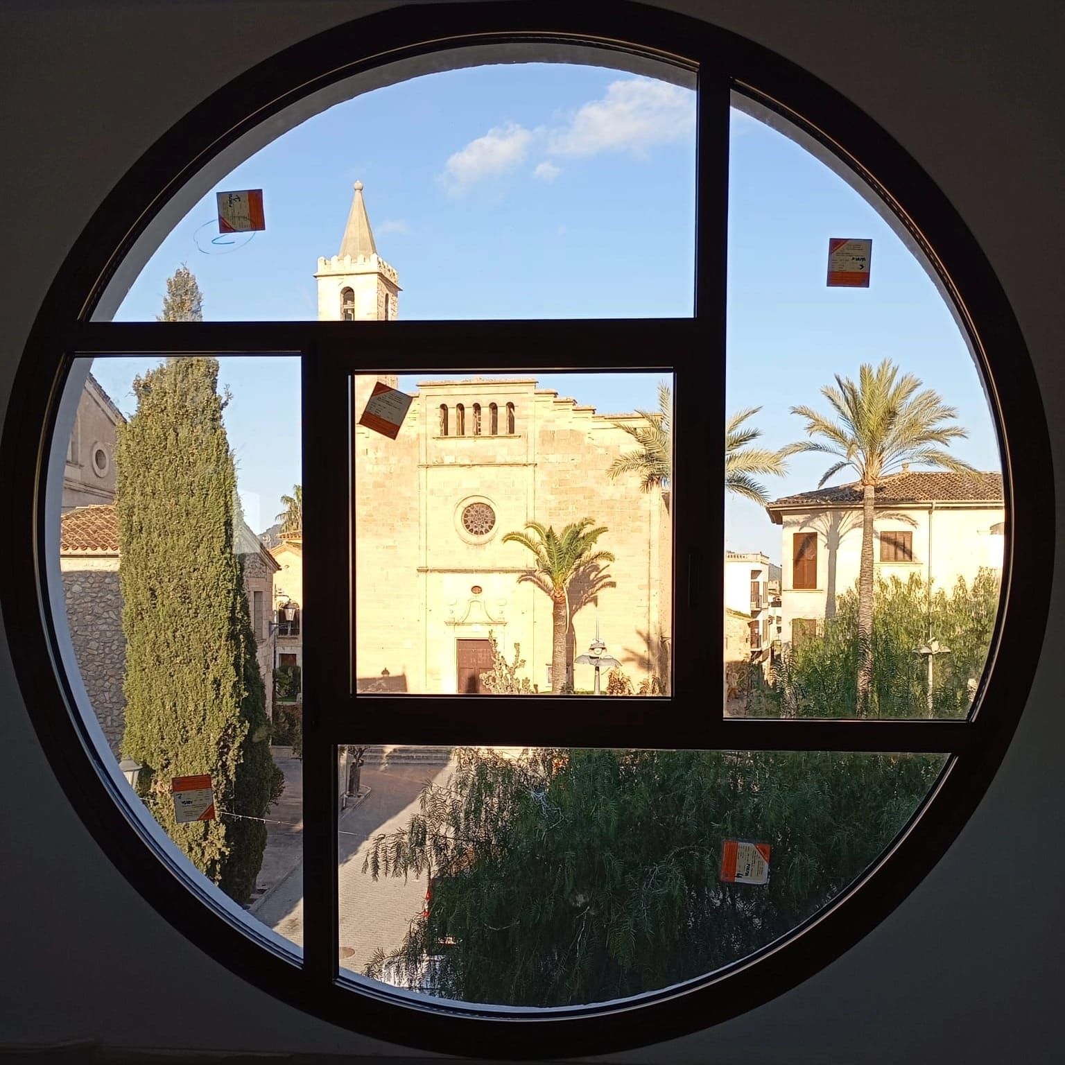 Una ventana circular enmarca una iglesia y un patio con palmeras y cielo azul.