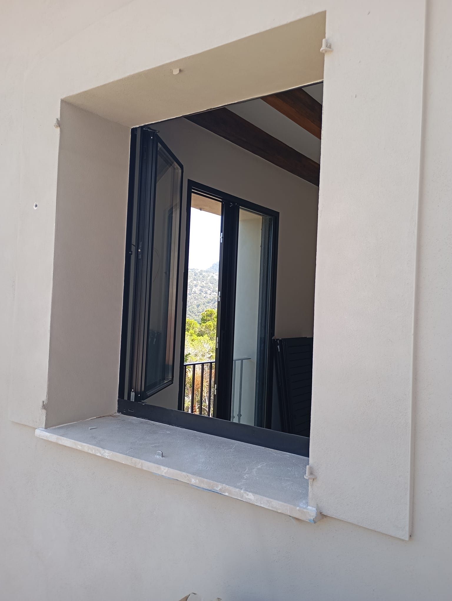 Ventana exterior con contraventanas negras abiertas, alféizar de piedra y pared exterior gris claro.