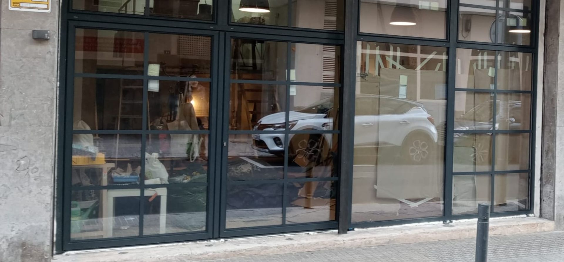 Escaparate con grandes ventanales que reflejan una escena callejera y un coche blanco aparcado. Marcos de ventana negros.