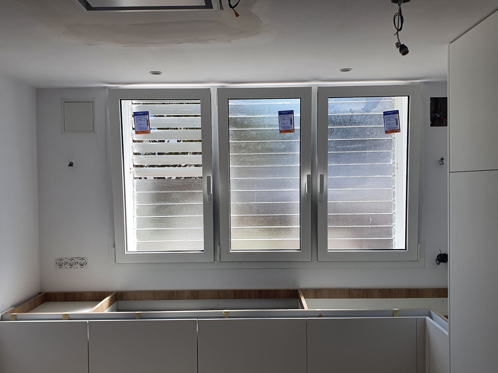 Tres ventanas blancas con persianas sobre gabinetes blancos en una cocina en construcción.