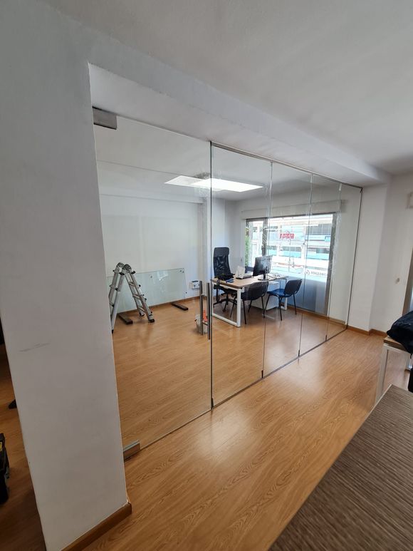 Oficina con paredes de vidrio, piso de madera, escritorio, silla y ventana.