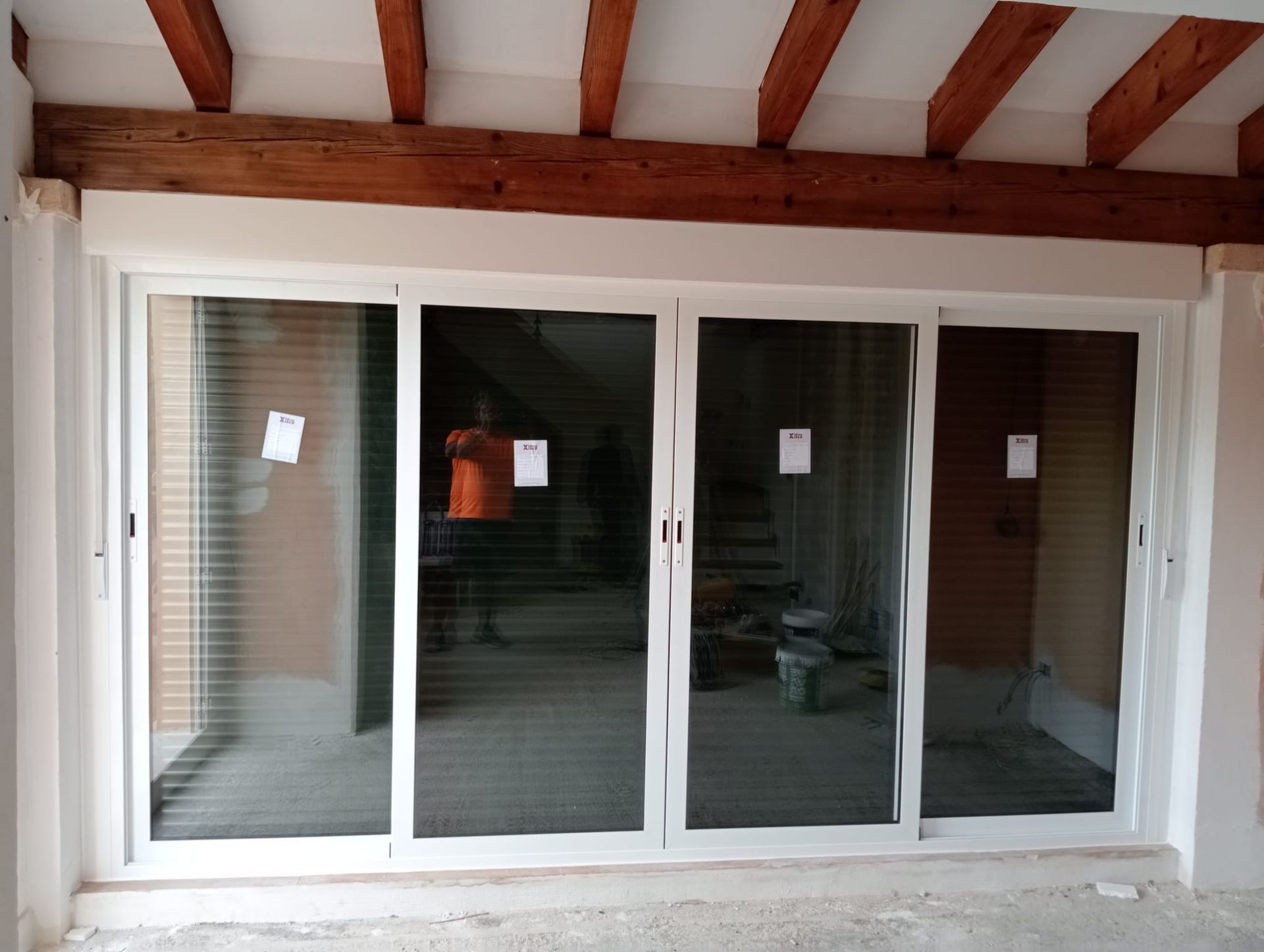 Puertas corredizas de vidrio con marcos blancos bajo vigas de madera, que reflejan una persona y una construcción interior.