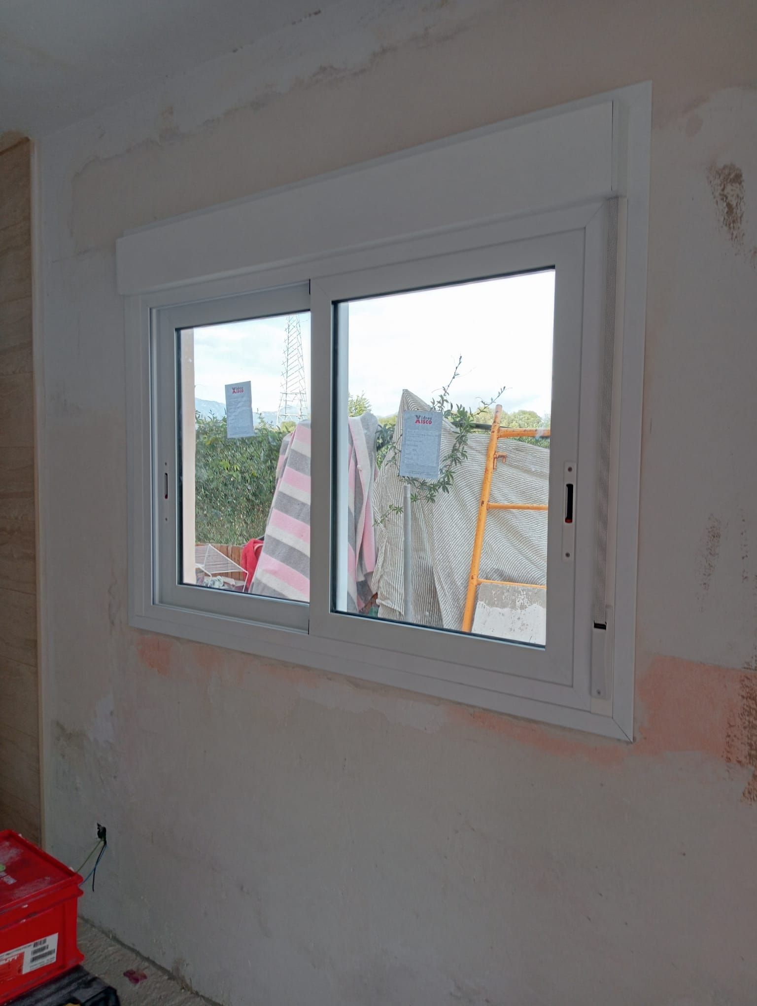 Ventana blanca con persiana incorporada en pared de yeso, con vista al exterior visible a través del vidrio.