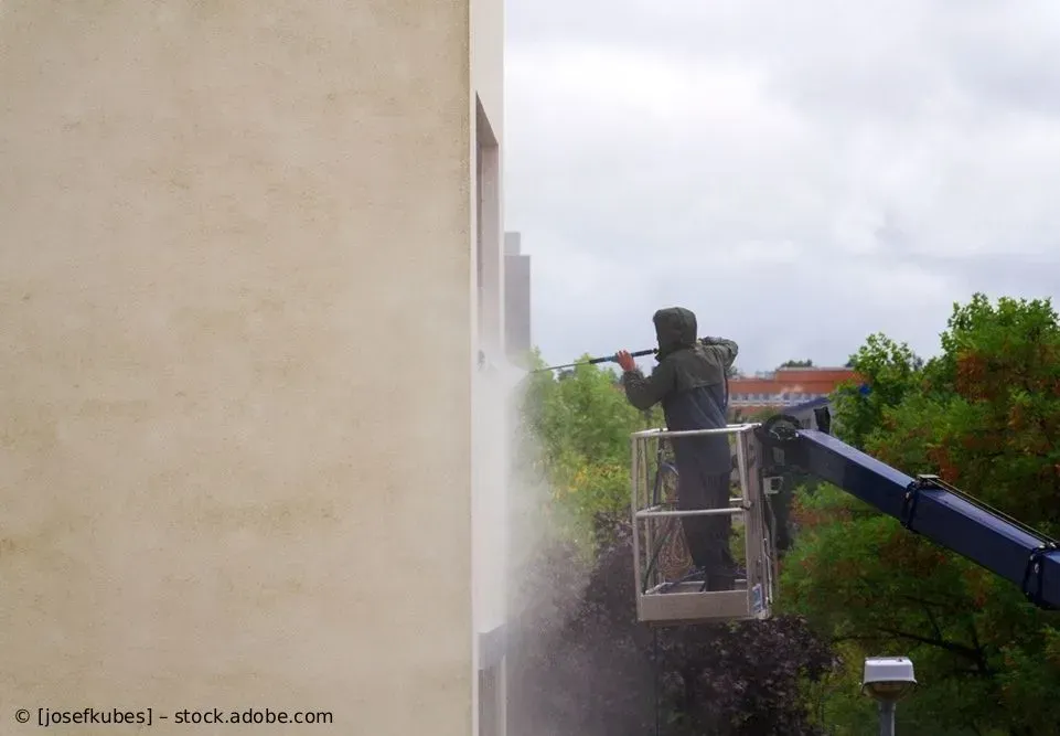 Ein Arbeiter besprüht auf einer Hebebühne die Seite eines beigen Gebäudes im Freien mit Wasser oder Farbe.