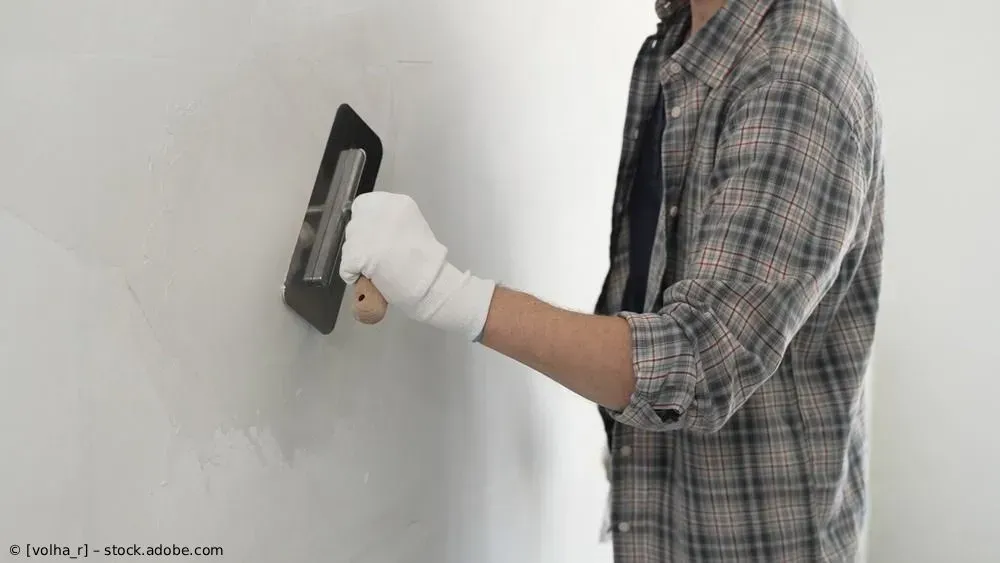 Eine Person in einem karierten Hemd bedient ein an der Wand montiertes Gerät, während sie einen weißen Gipsverband am Handgelenk trägt.