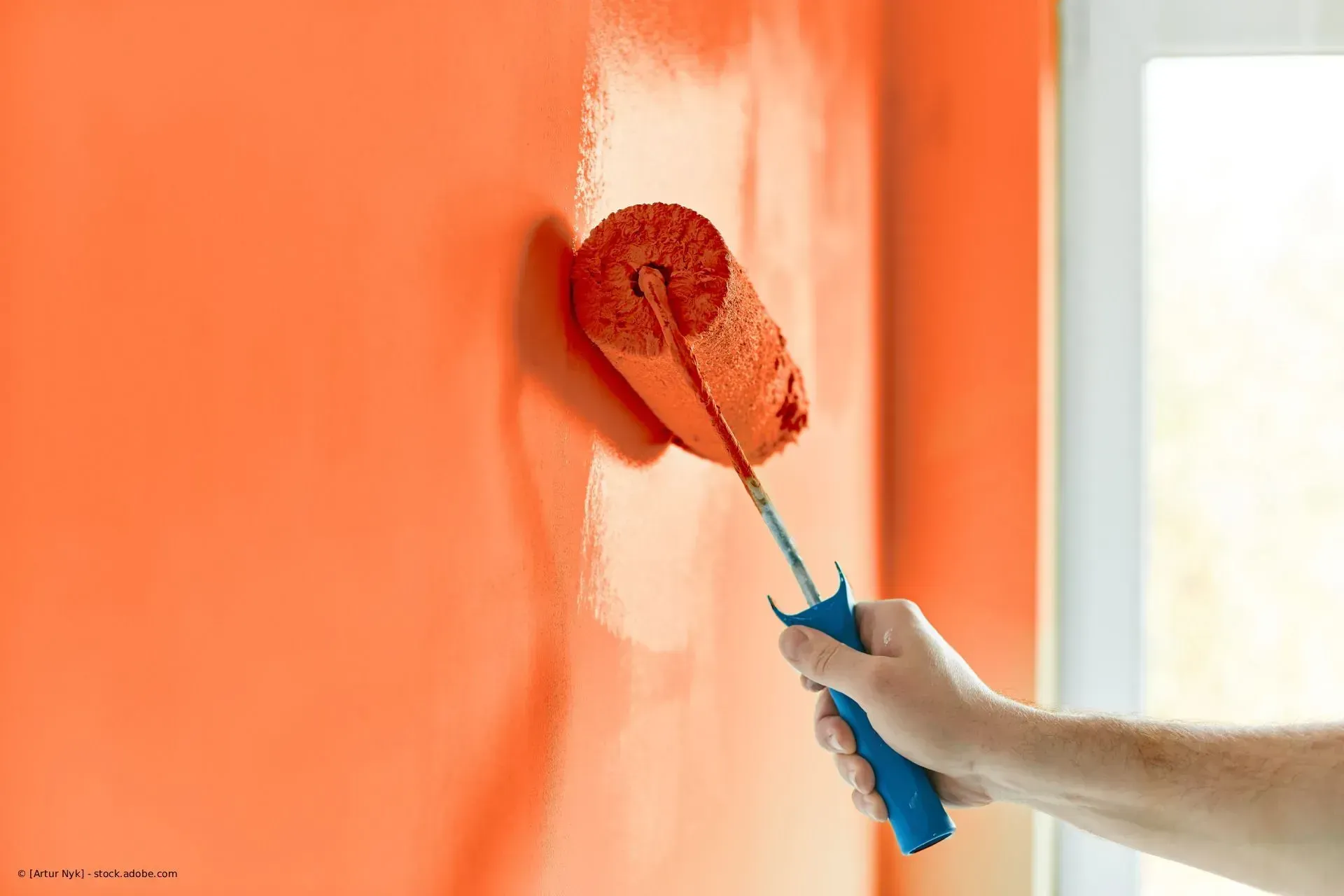 Eine orange Wand mit der Handfarbe und einer Farbrolle an einem blauen Griff streichen