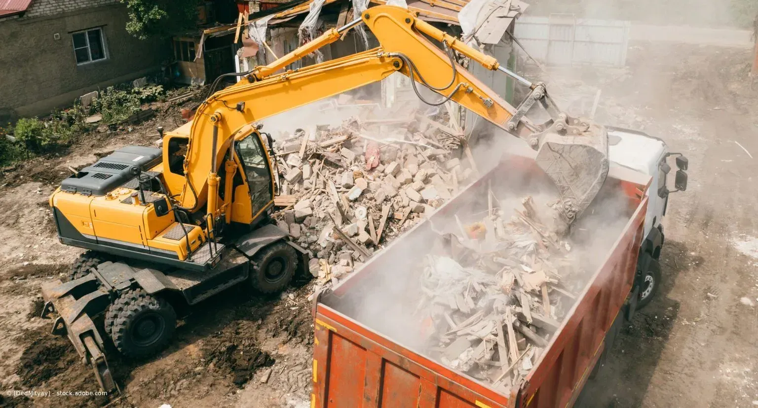 Ein gelber Bagger reißt Beton in einen orangefarbenen Container und wirbelt dabei Staub auf einer Baustelle auf.