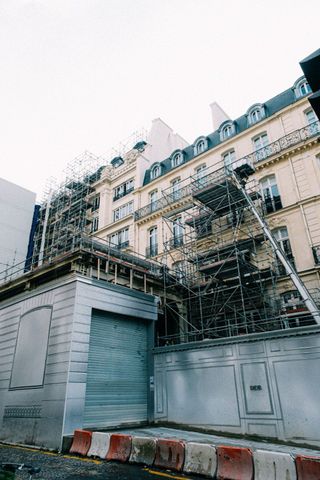 Ein Baugerüst verdeckt die beige Fassade eines Gebäudes neben einem grauen Tor und rot-weißen Absperrungen an einer Straße.