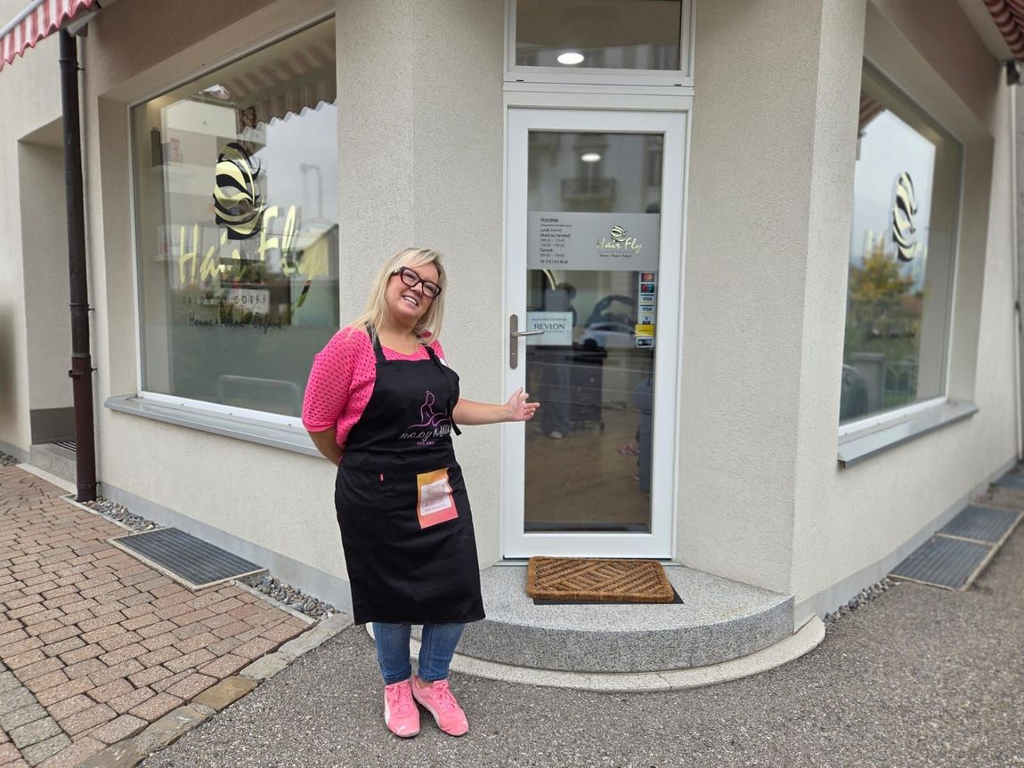 Une femme en tablier se tient devant l'entrée d'un magasin. Elle sourit, près des vitrines. Vue extérieure.