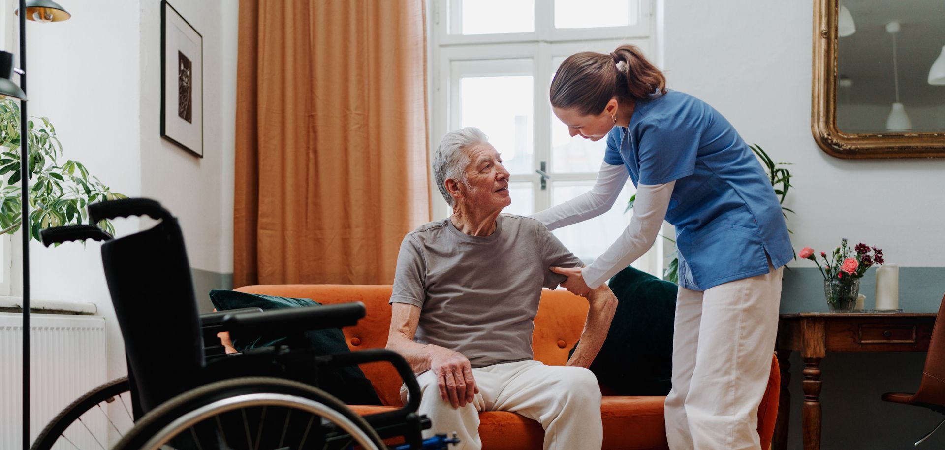 Eine Krankenschwester hilft einem älteren Mann im Rollstuhl, sich auf eine Couch zu setzen.
