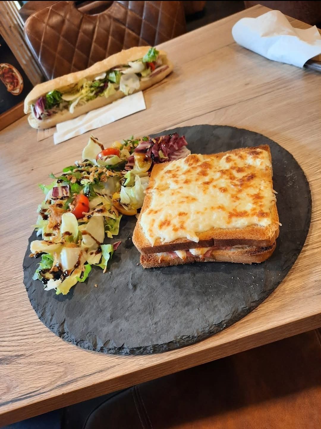 Croque-monsieur avec salade sur une assiette en ardoise, sandwich baguette en arrière-plan, table en bois.