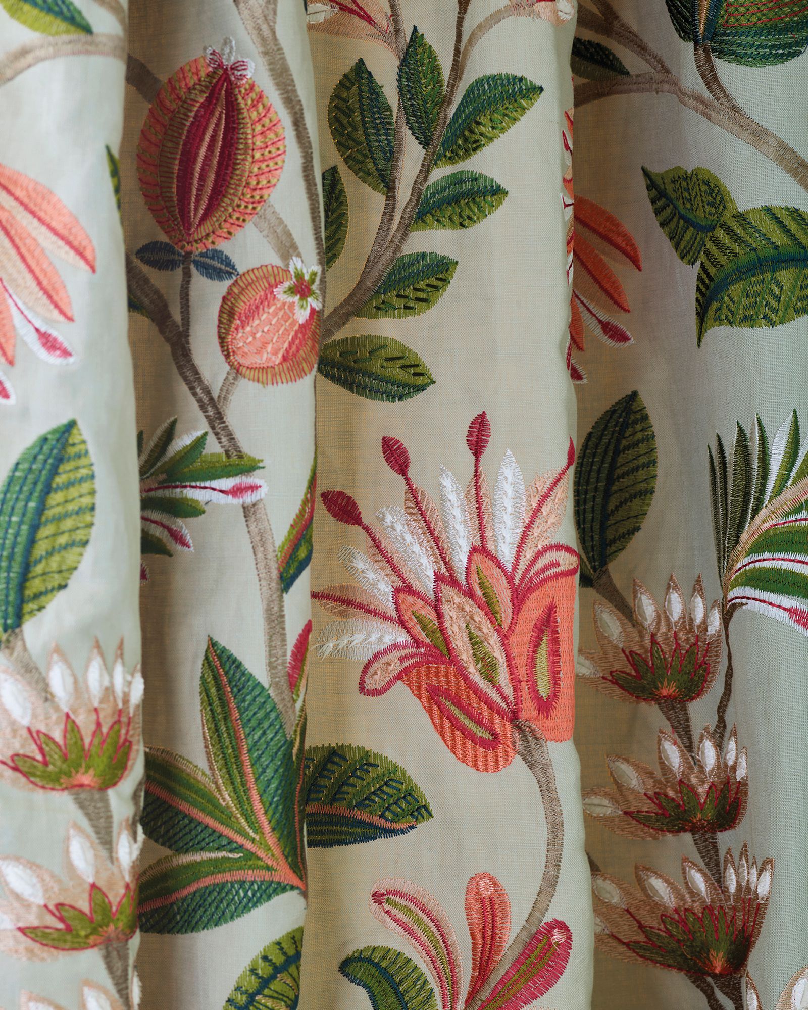 Bestickter Stoff mit Blumen- und Blattmustern in Orange, Grün und Weiß auf hellbeigem Hintergrund.