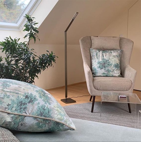 Gemütliche Leseecke mit Sessel, gemusterten Kissen, Stehlampe und kleinem Tisch in der Nähe eines Fensters und einer Pflanze.