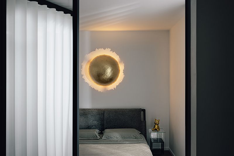 Schlafzimmereinrichtung mit goldener Wandleuchte über dem Bett; Fenster mit weißen Vorhängen auf der linken Seite.