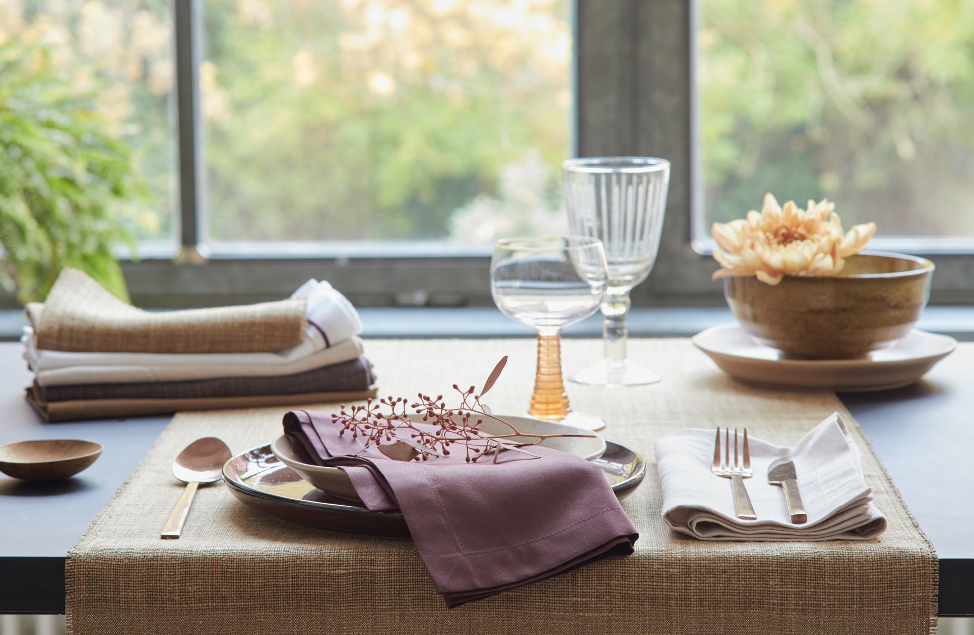Tischgedeck mit Tellern, Besteck, Gläsern, Servietten und einem Blumenarrangement vor einem Fenster.