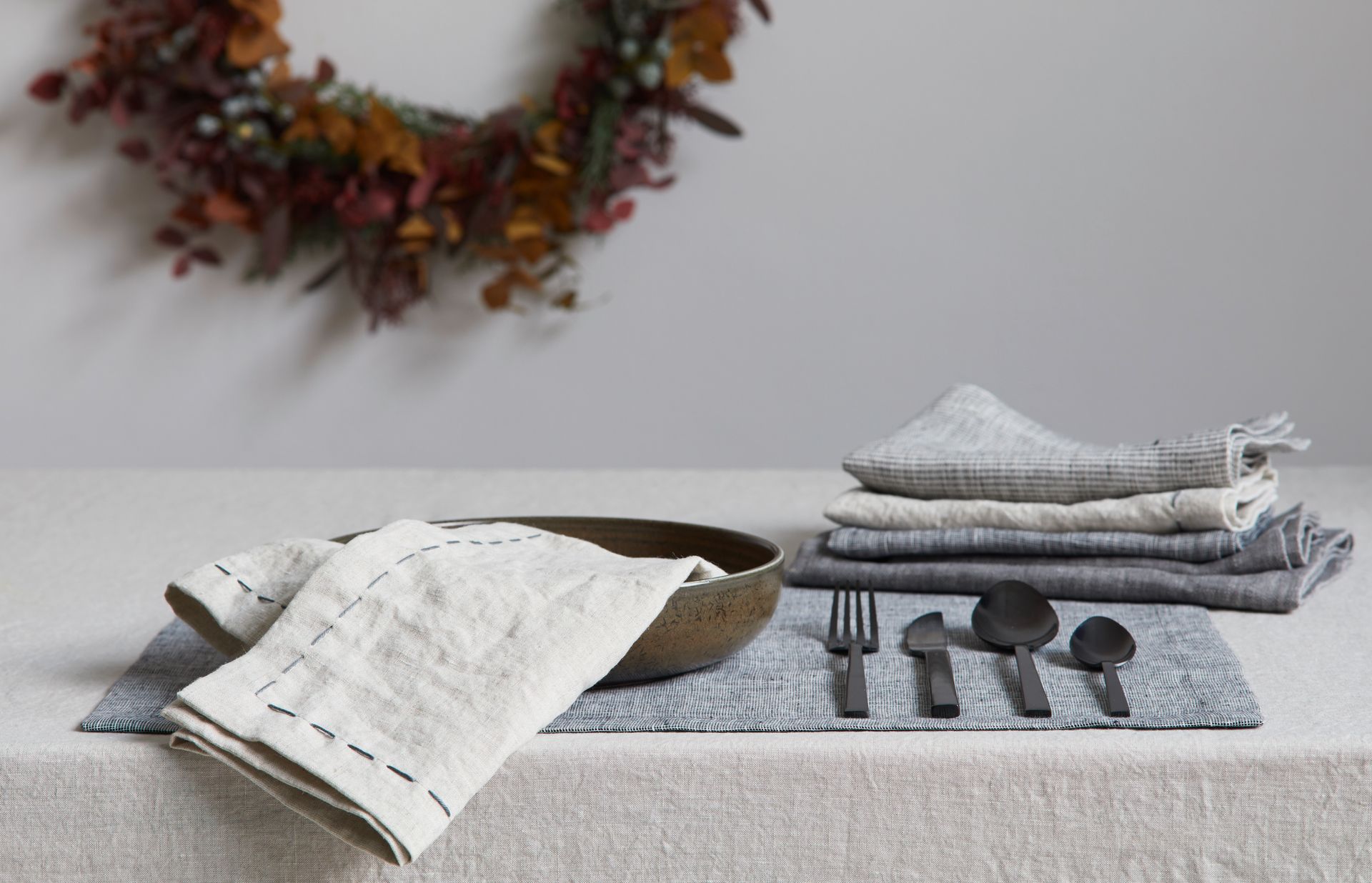 Tischgedeck mit Leinenservietten, schwarzem Besteck, einer Schüssel und einem Kranz.