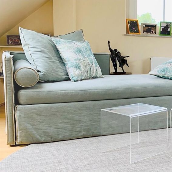 Eine hellblaue Chaiselongue mit dekorativen Kissen steht auf einem Teppich neben durchsichtigen Beistelltischen.