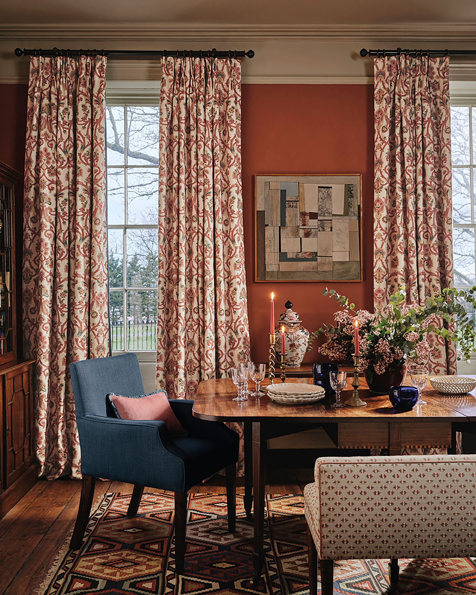Esszimmer mit gemusterten Vorhängen, orangefarbener Wand, gedecktem Holztisch für das Abendessen, blauen Stühlen und einem abstrakten Gemälde.