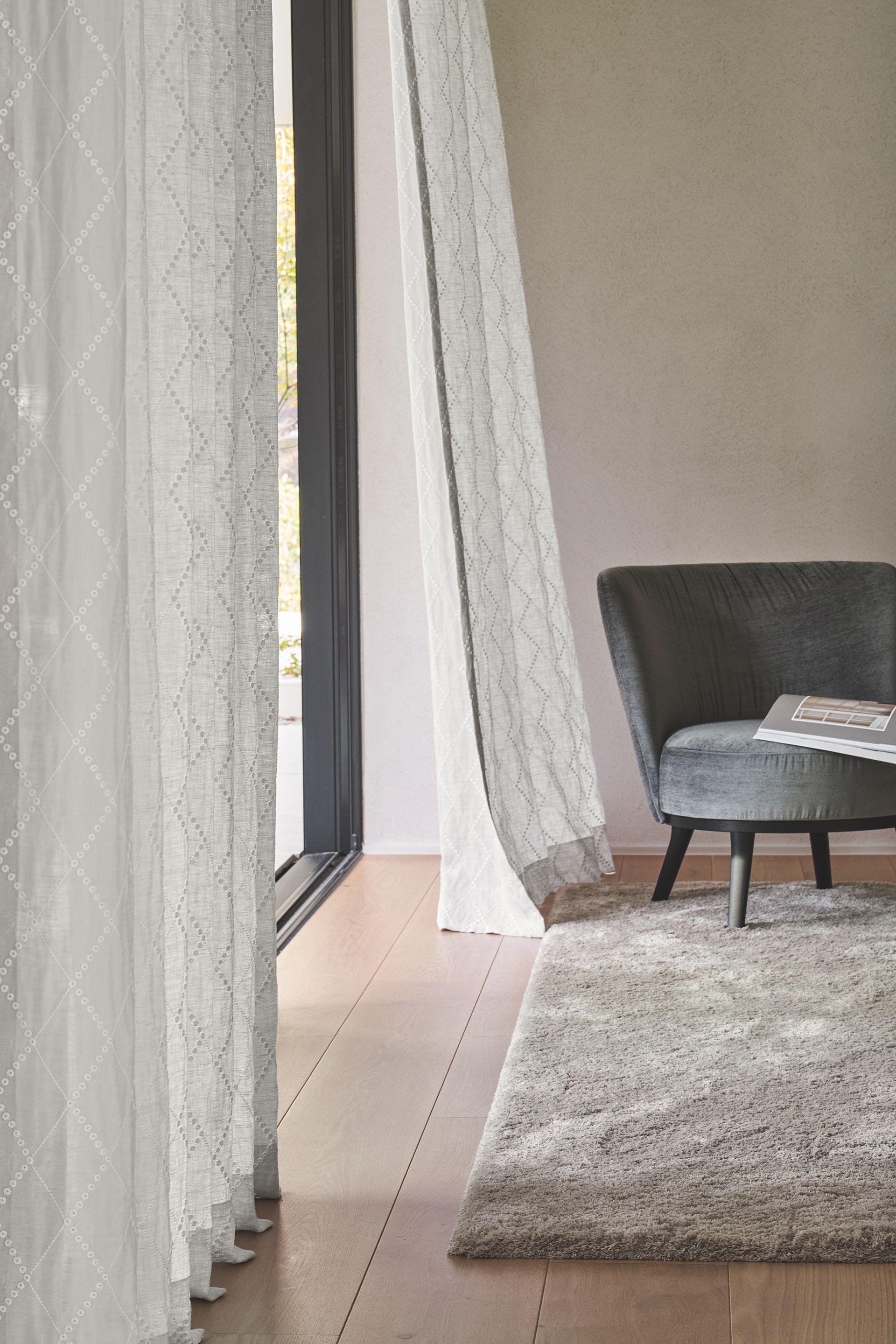 Transparente weiße Vorhänge rahmen einen Türrahmen ein, auf einem Holzboden stehen ein grauer Sessel und ein zotteliger Teppich.
