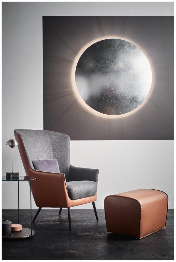 Gemütliche Wohnzimmerszene: Grauer Sessel, Lederhocker, kreisförmiges Kunstwerk mit hell- und dunkelgrauer Wand.