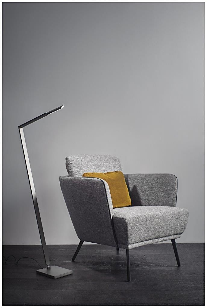 Silberne Stehlampe neben einem grauen Sessel mit einem gelben Kissen vor einer grauen Wand.