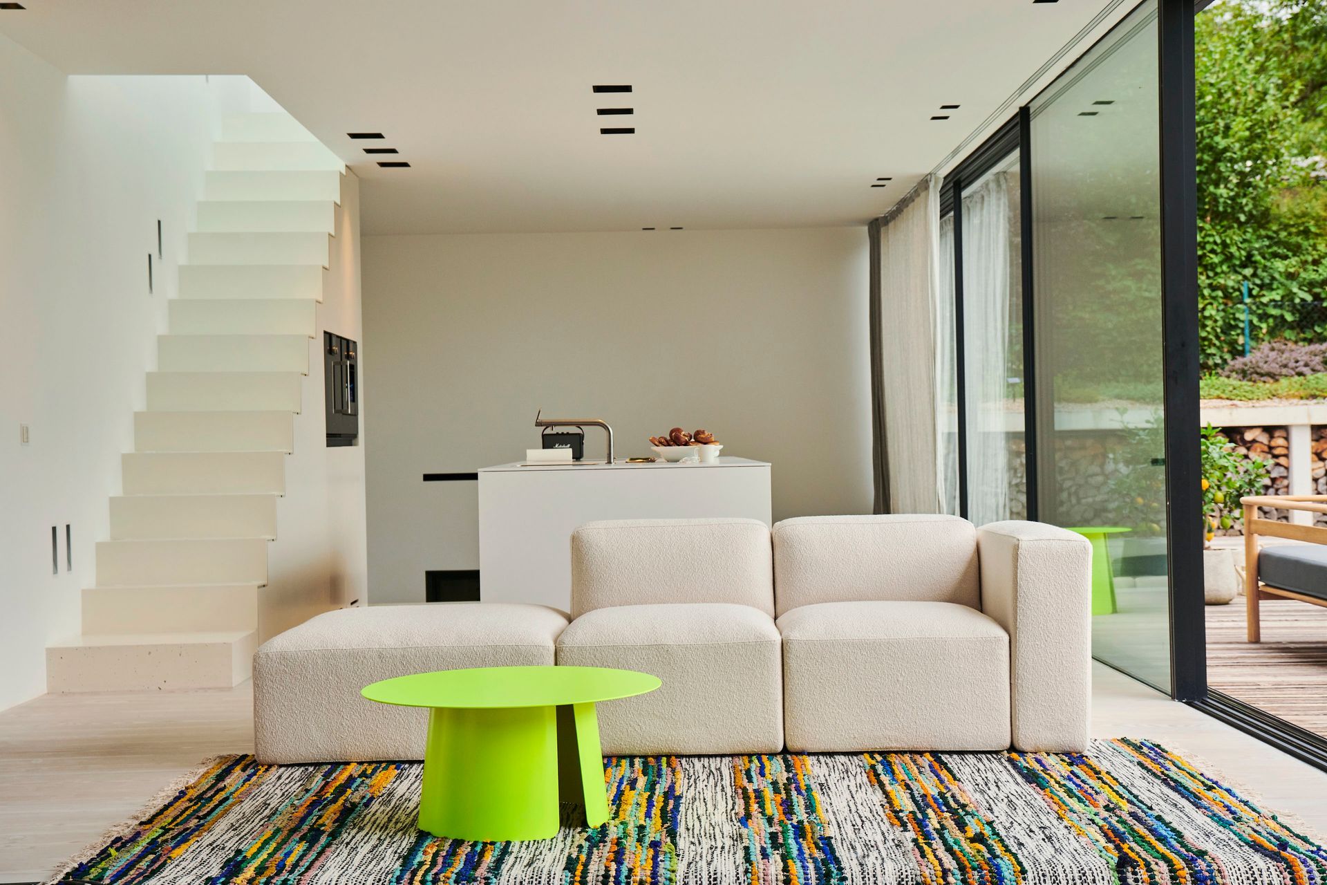 Wohnzimmer mit weißem Sofa, grünem Tisch, buntem Teppich und Treppe. Glasschiebetür zu einer Terrasse.