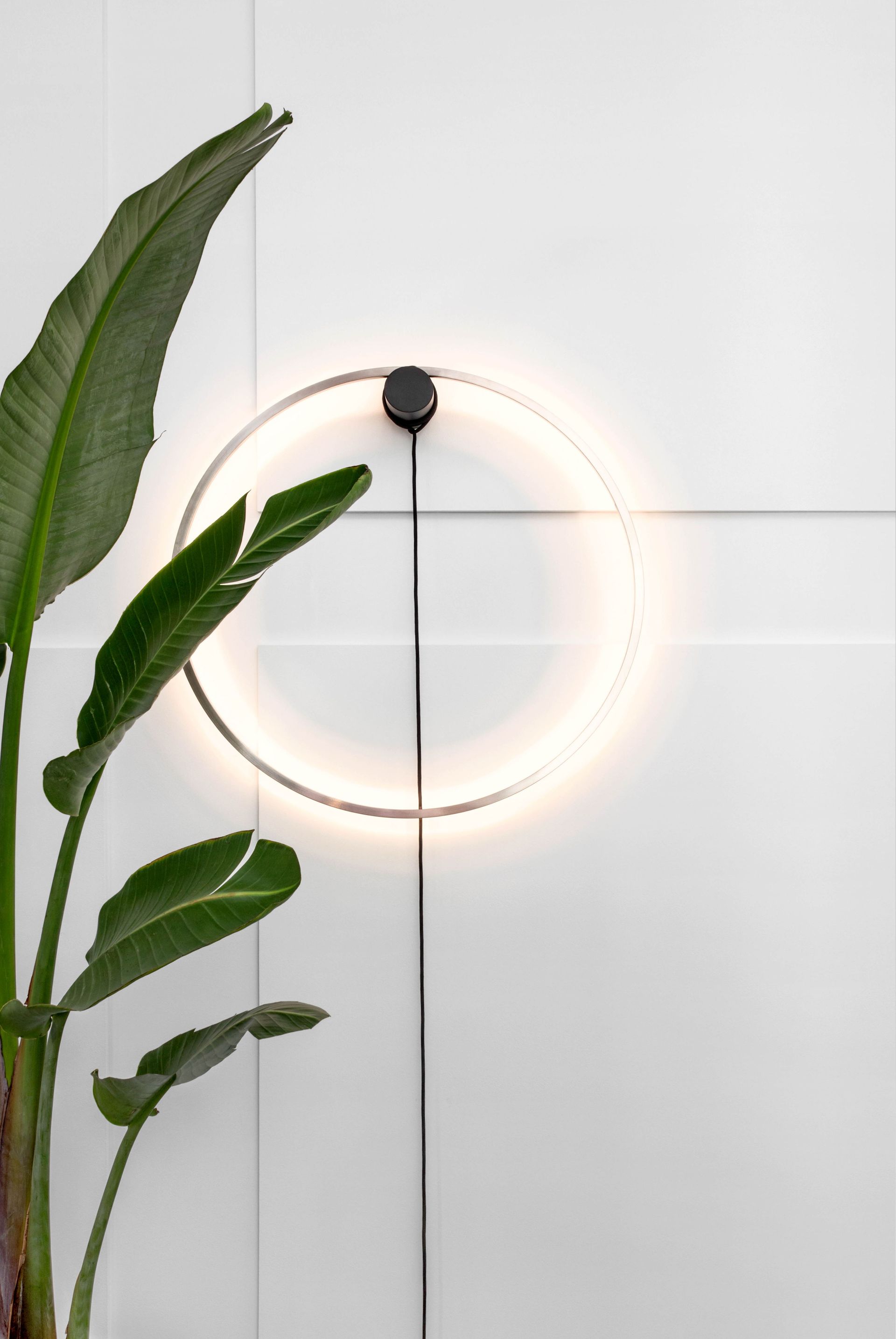 Eine moderne runde Wandleuchte beleuchtet eine weiß geflieste Wand mit einer Topfpflanze.