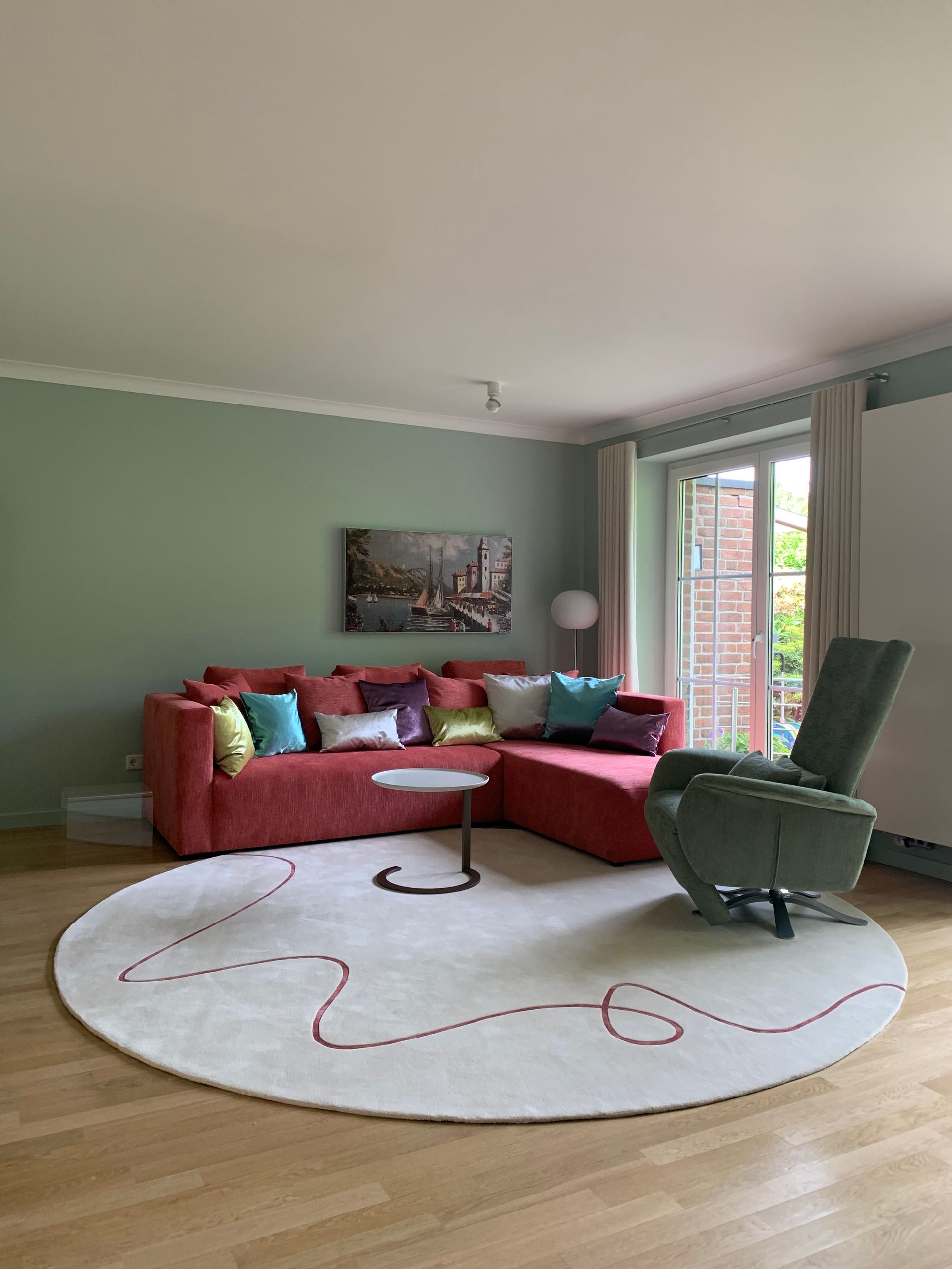 Wohnzimmer mit roter Sofagarnitur, grünem Ruhesessel, rundem Teppich und grünen Wänden.