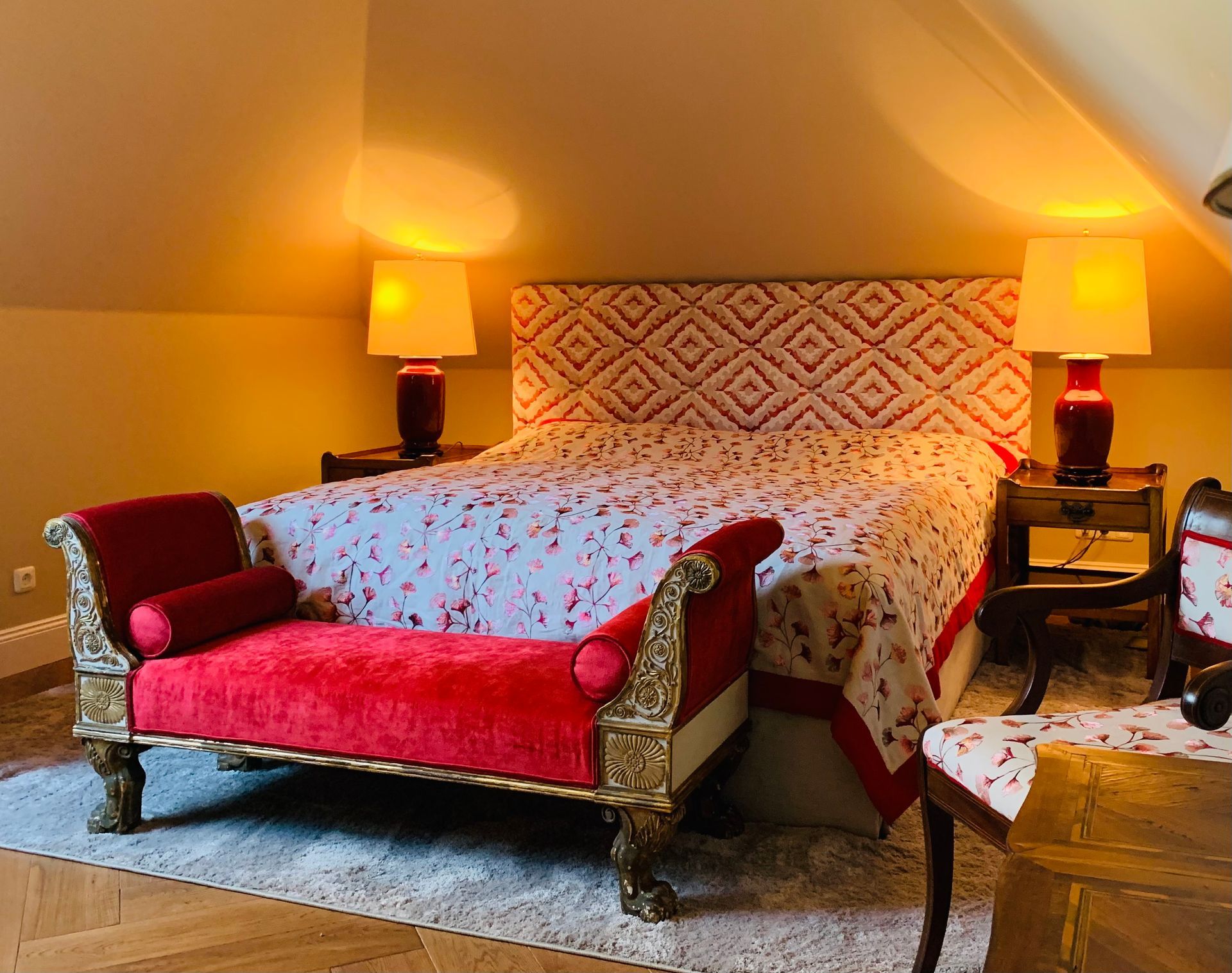 Gemütliches Schlafzimmer mit roter Chaiselongue, gemustertem Bett und Nachttischlampen.