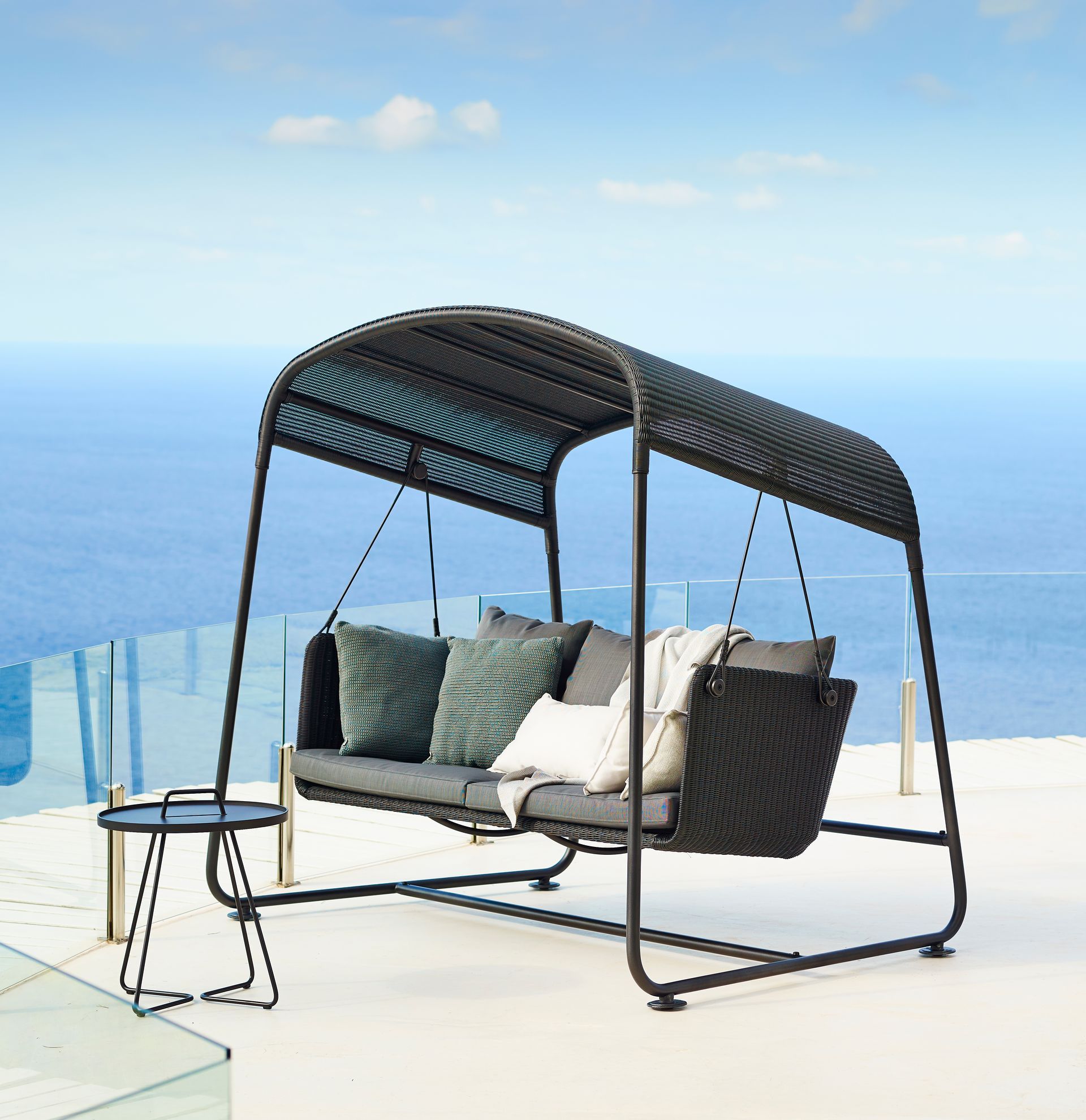 Schwarze Gartenschaukel mit Baldachin und Kissen, neben einem kleinen Tisch, mit Blick auf den Ozean.