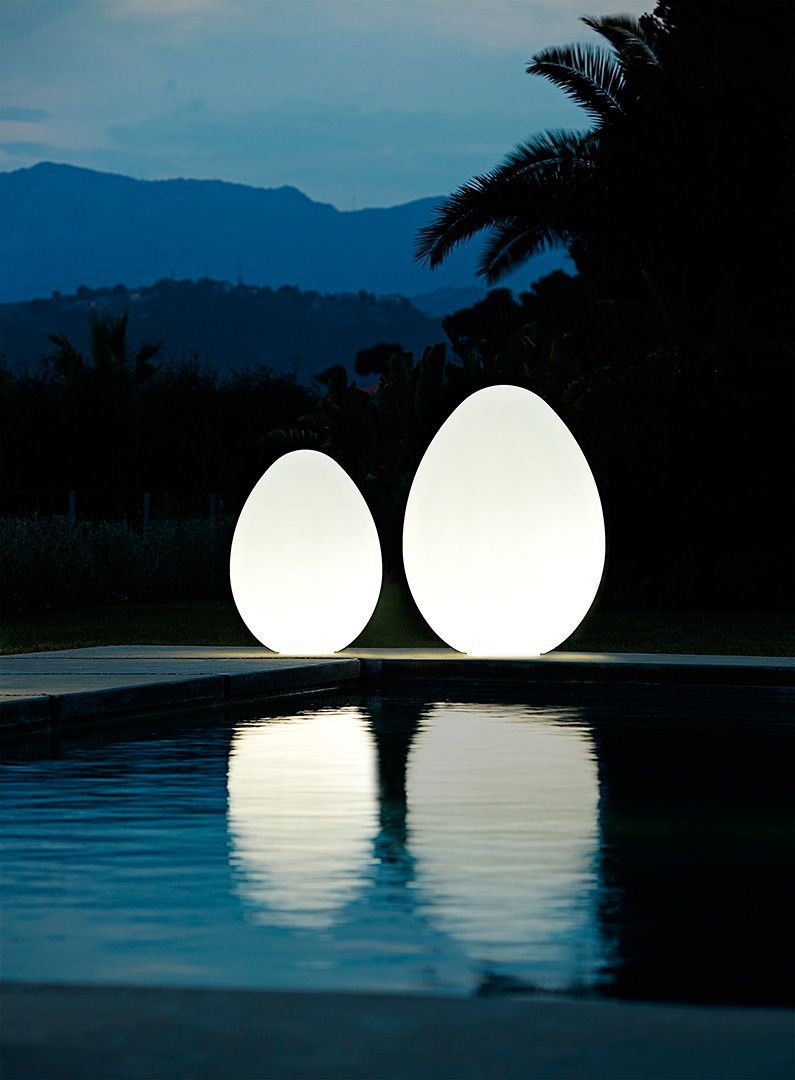 Zwei leuchtende, eiförmige Lichter spiegeln sich in der Abenddämmerung in einem Teich, im Hintergrund sind Berge zu sehen.