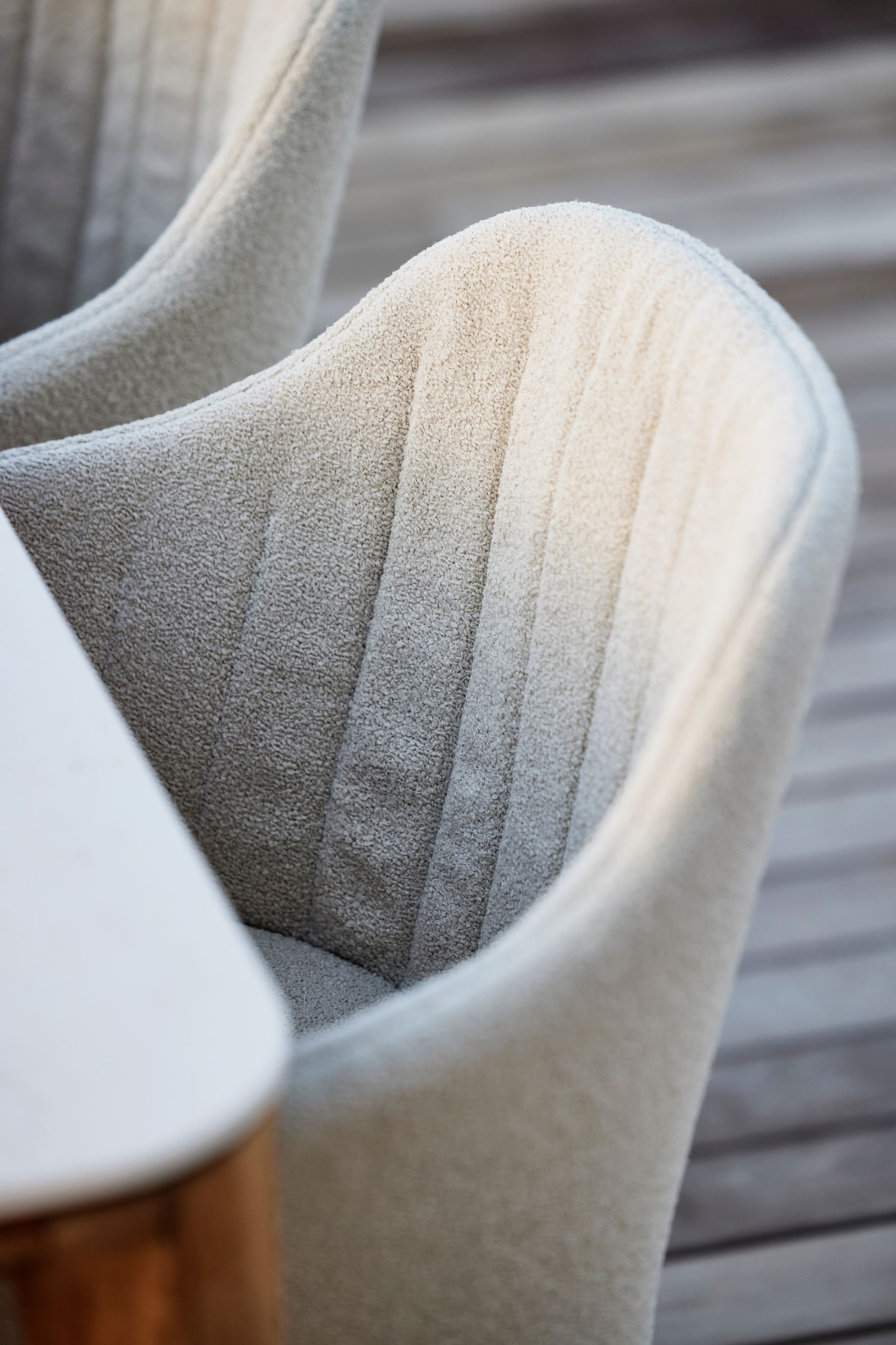 Nahaufnahme einer hellgrauen, strukturierten Stuhllehne mit vertikalen Nahtdetails neben einer weißen Tischkante.