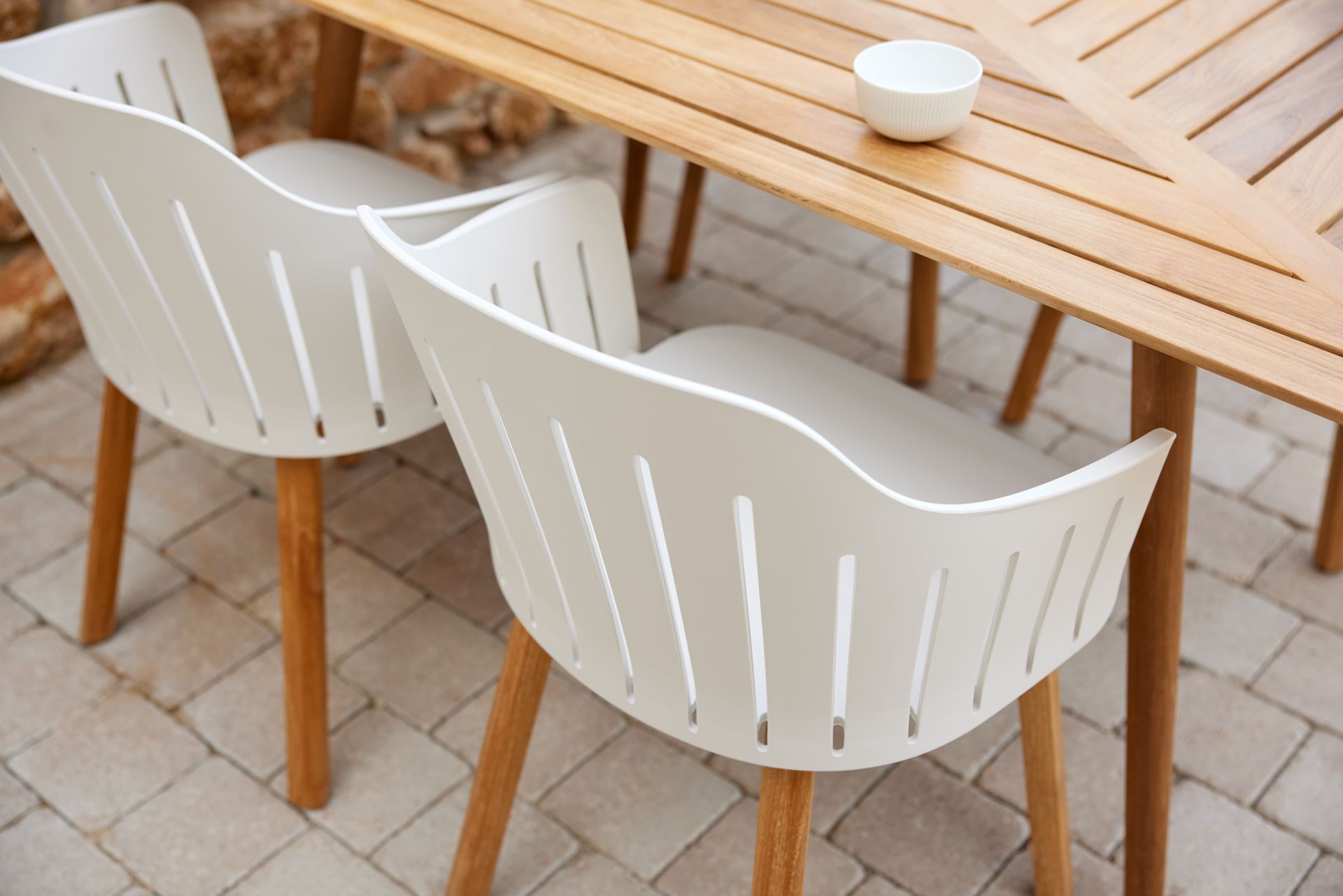Zwei weiße Stühle mit Holzbeinen und ein Holztisch auf einer Steinterrasse, mit einer weißen Schüssel.