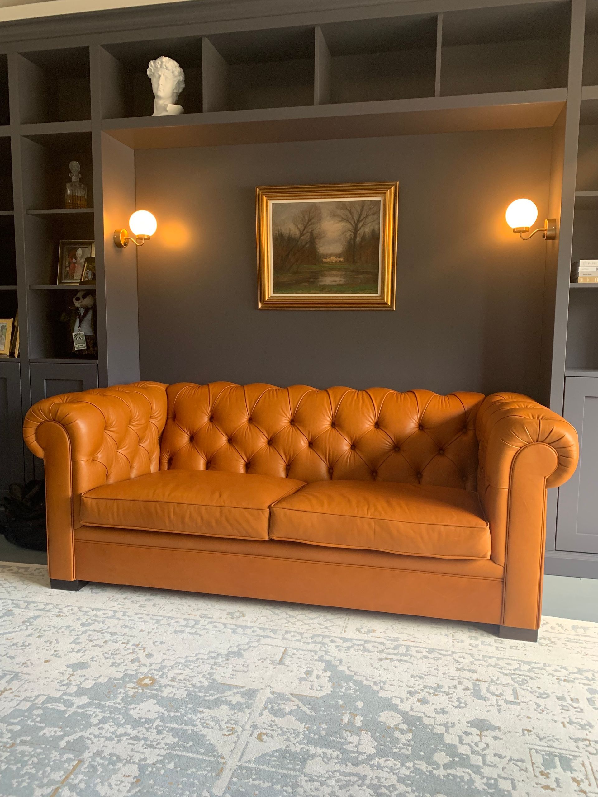 Orangefarbenes Ledersofa mit Polsterknöpfen vor einer dunkelgrauen Wand mit Wandlampen und Kunstwerken.