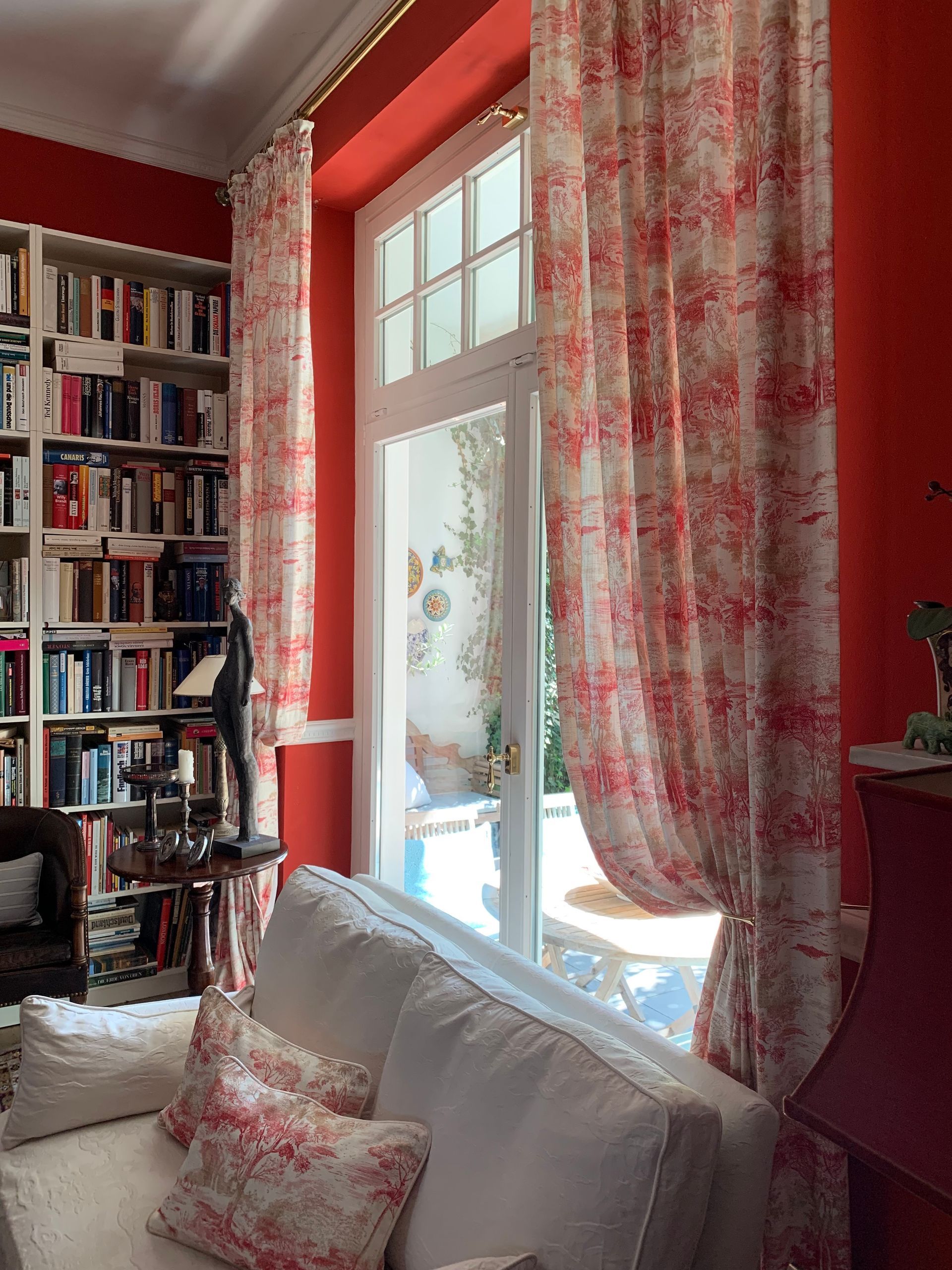 Ein roter Raum mit weißem Sofa, Bücherregalen und rot gemusterten Vorhängen, die ein Fenster zu einer Außenterrasse einrahmen.