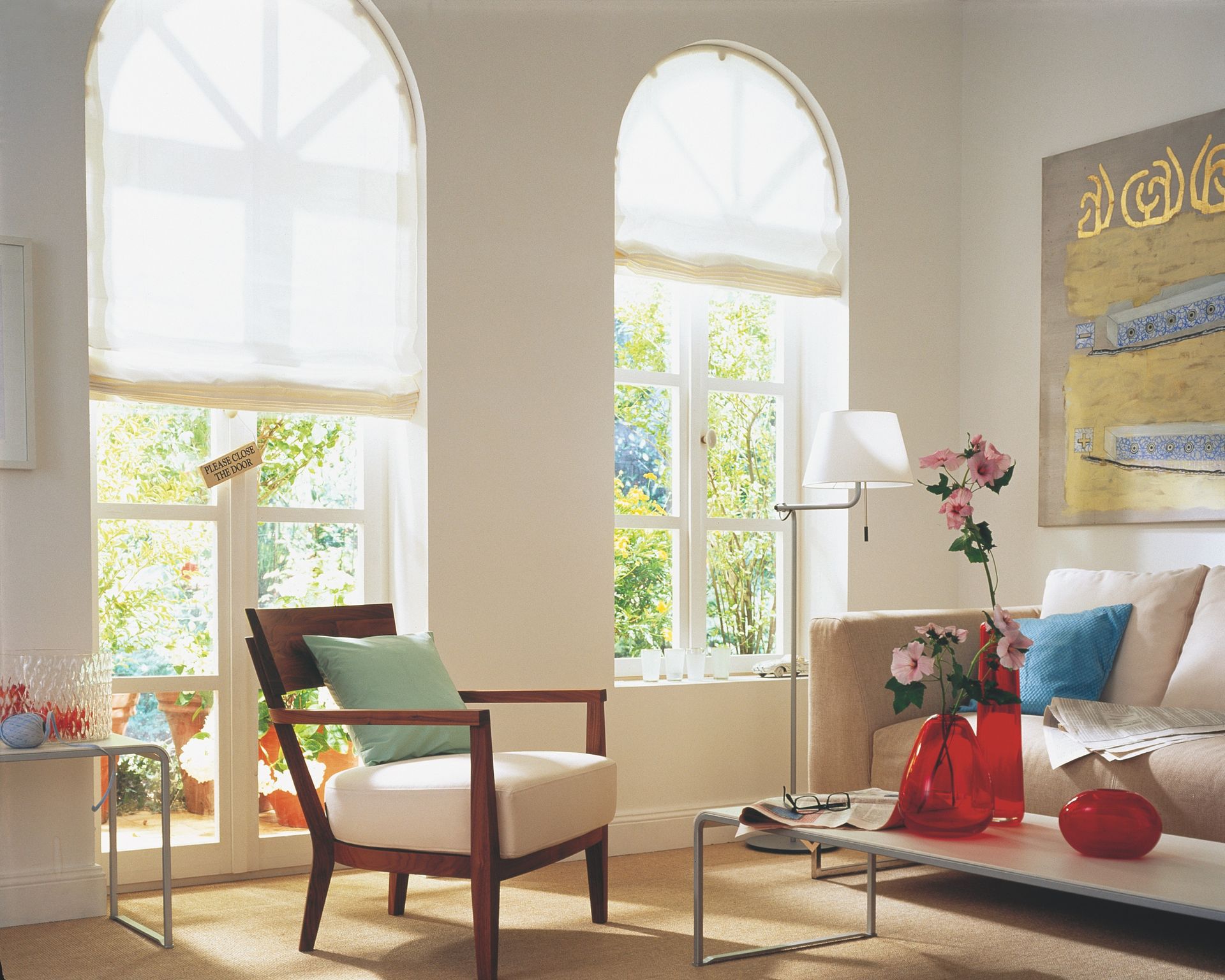 Wohnzimmer mit Bogenfenstern, die sich zum Garten hin öffnen. Ein Stuhl, ein Sofa, ein Tisch und eine Lampe sind vorhanden.