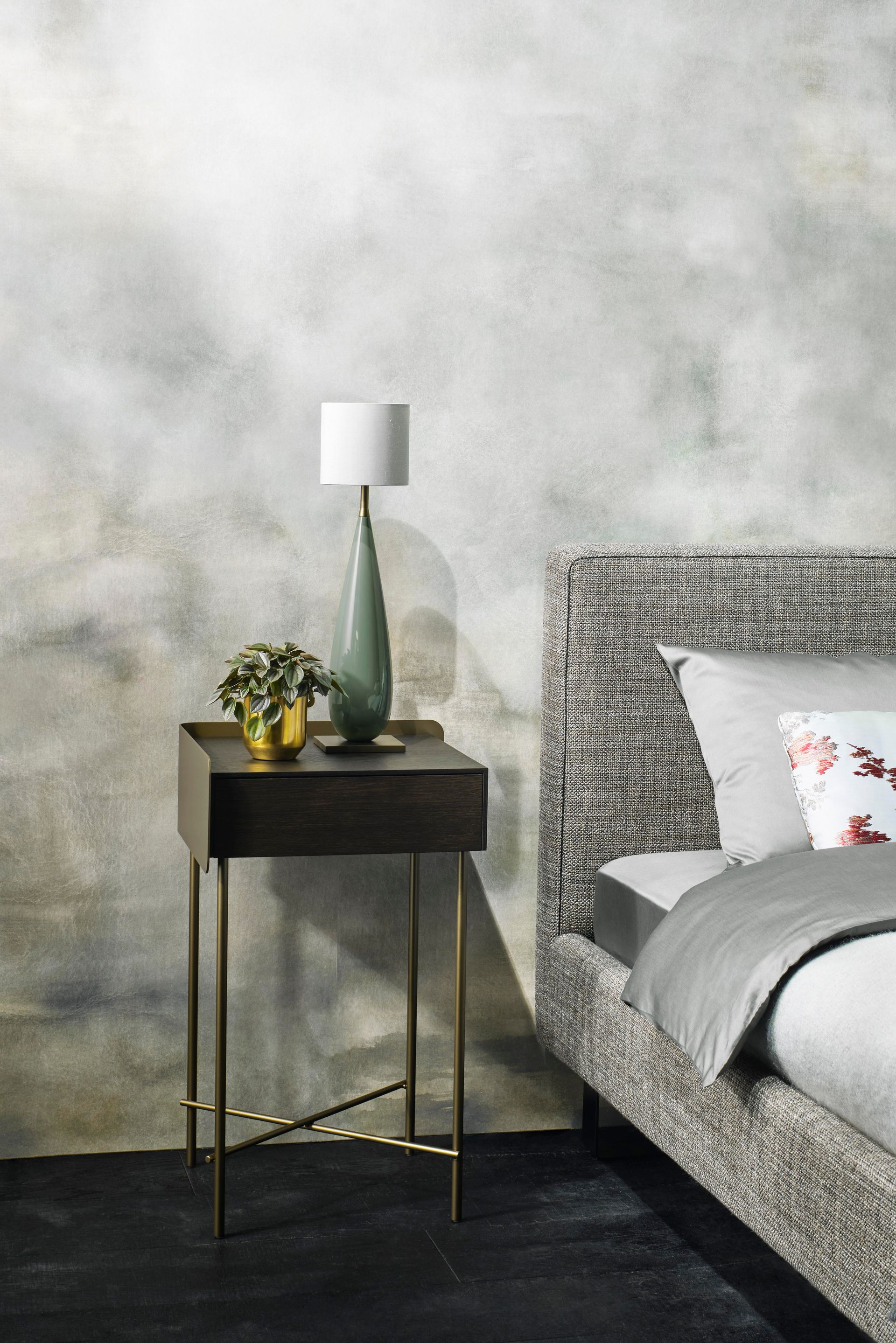 Nachttisch mit Lampe und Pflanze neben einem gemusterten Bett vor einer strukturierten grauen Wand.
