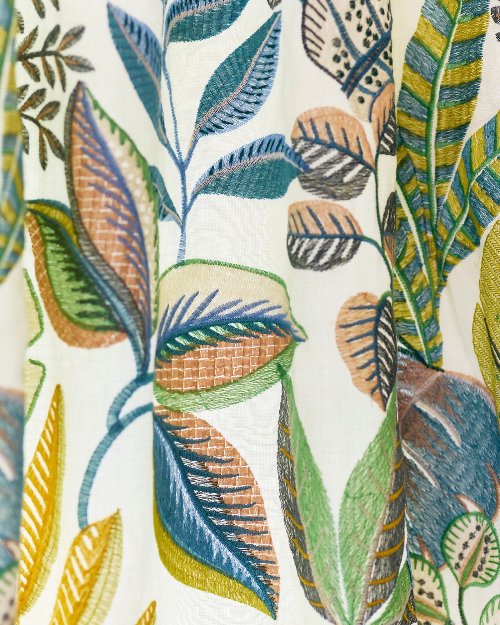 Nahaufnahme eines weißen Stoffes mit bunt gesticktem botanischem Muster aus Blättern und Stängeln in Grün-, Blau-, Braun- und Gelbtönen.