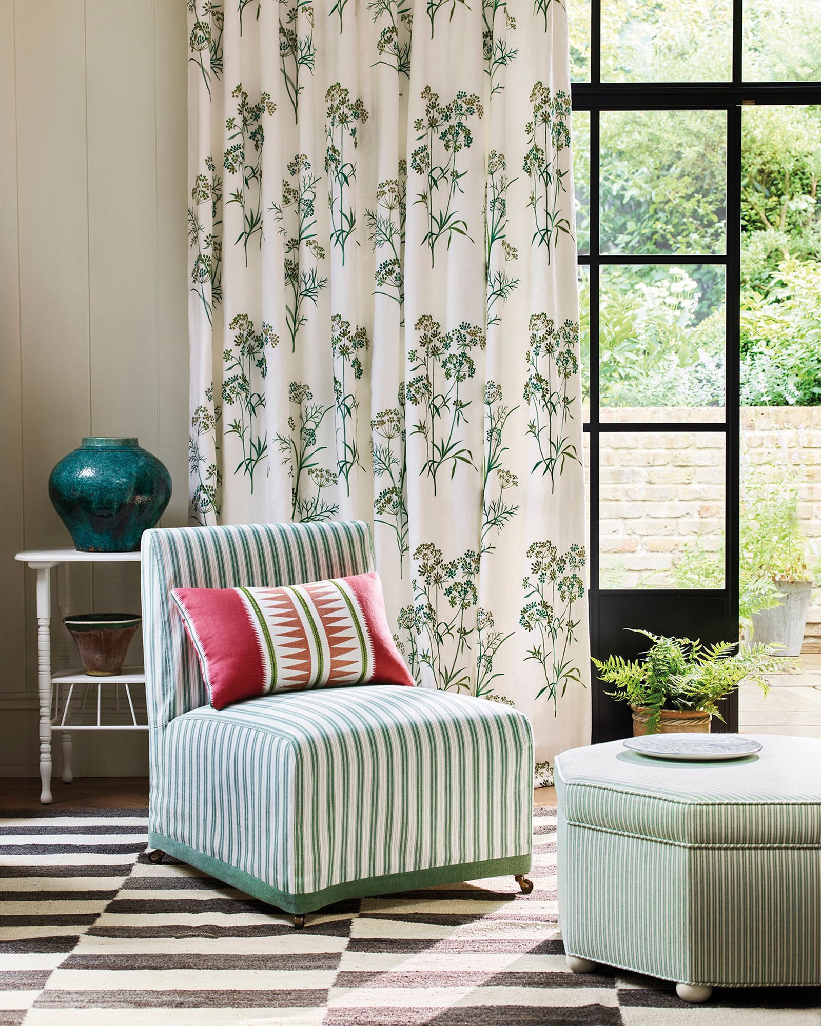 Ein Zimmer mit einem gestreiften Stuhl, geblümten Vorhängen und einem Blick nach draußen durch ein schwarz gerahmtes Fenster.