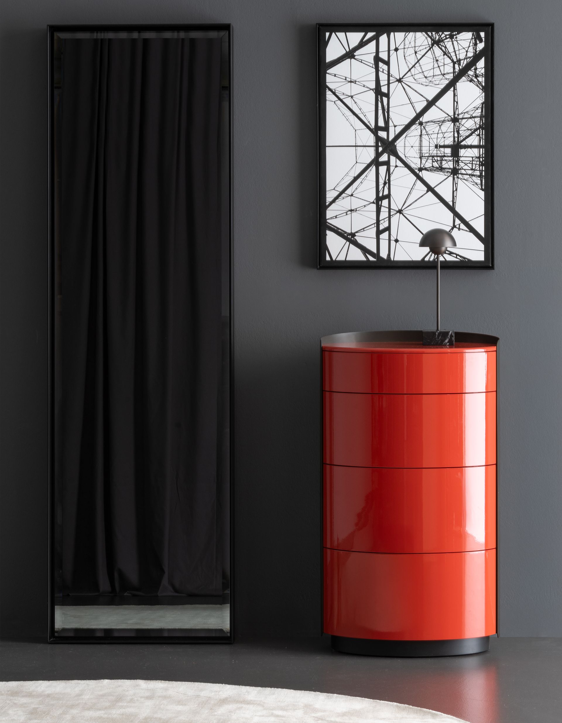 Roter, runder Schrank mit Kunst, einem schwarzen Spiegel und einer kleinen Lampe vor einer dunkelgrauen Wand.