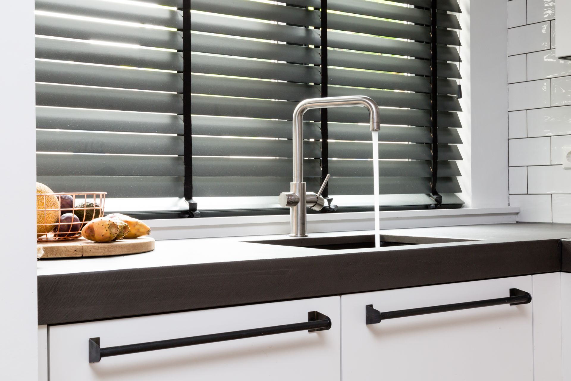 Küchenspüle mit fließendem Wasser, dunkle Arbeitsplatte, weiße Schränke mit schwarzen Griffen und ein Fenster mit Jalousien.
